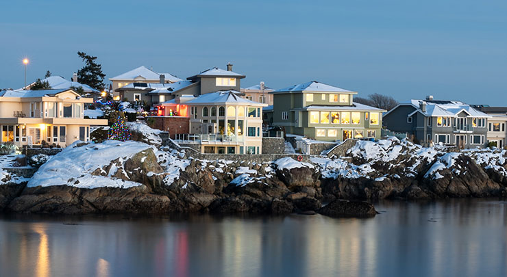 Snow covered coastal homes at dusk. Light reflects on the water in the foreground.